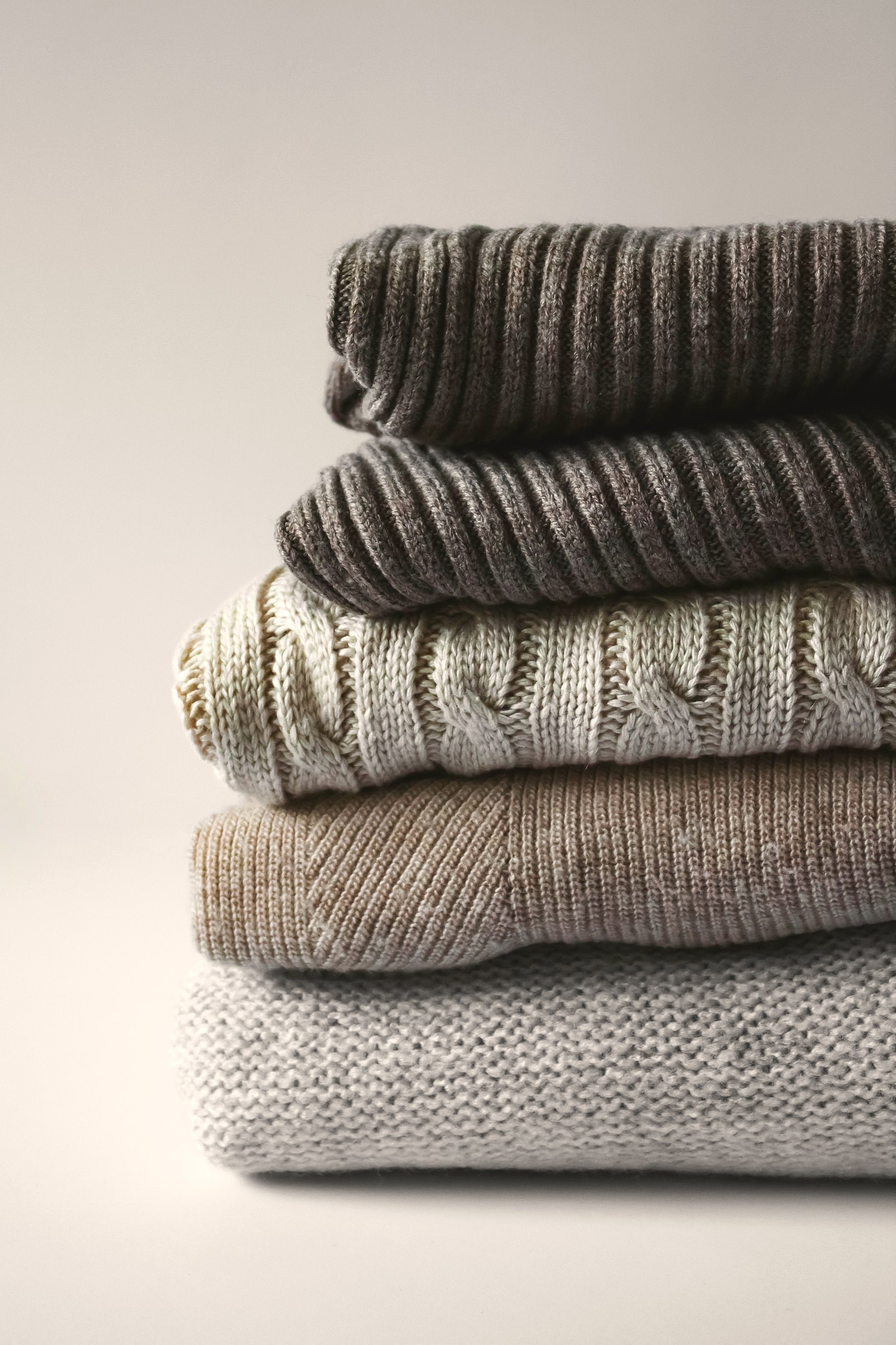 A folded stack of knits in charcoal, grey and stone, ribs and cables visible