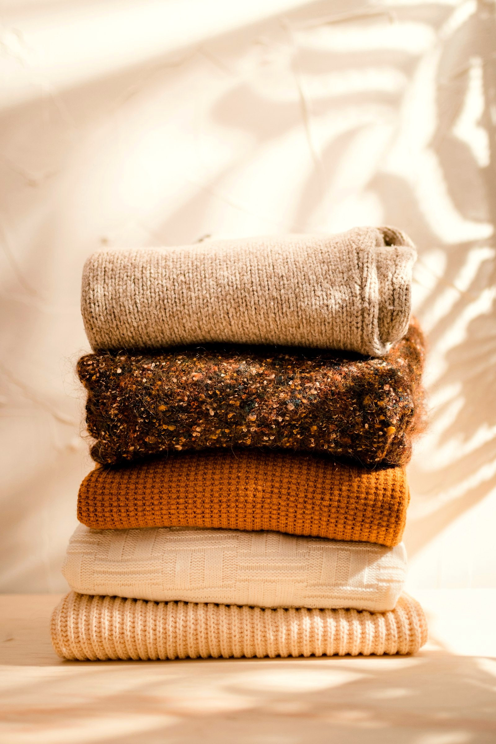 A stack of folded knits in oatmeal, rust, ochre and cream in sunlight with leaf shadows
