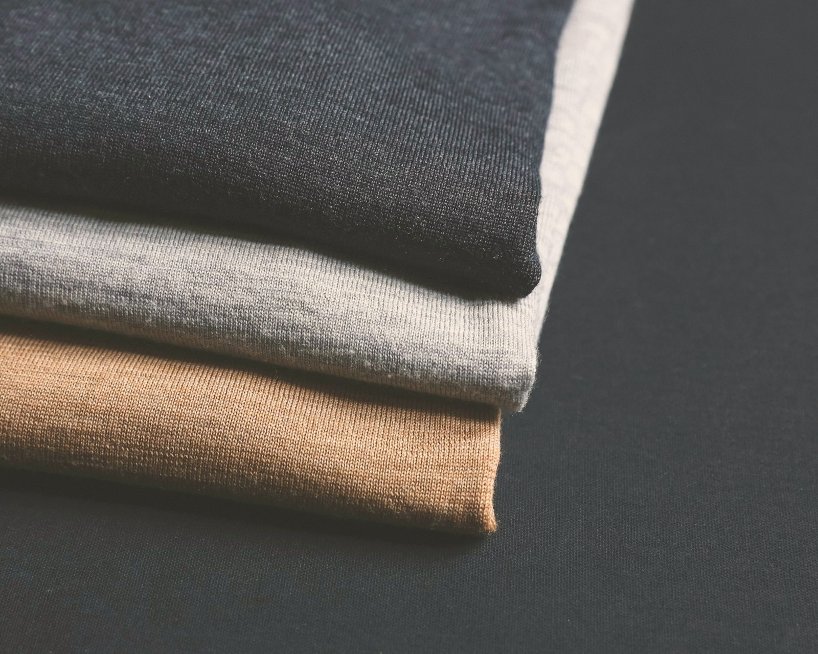 Three folded fine knits in navy, grey and camel on a dark slate surface