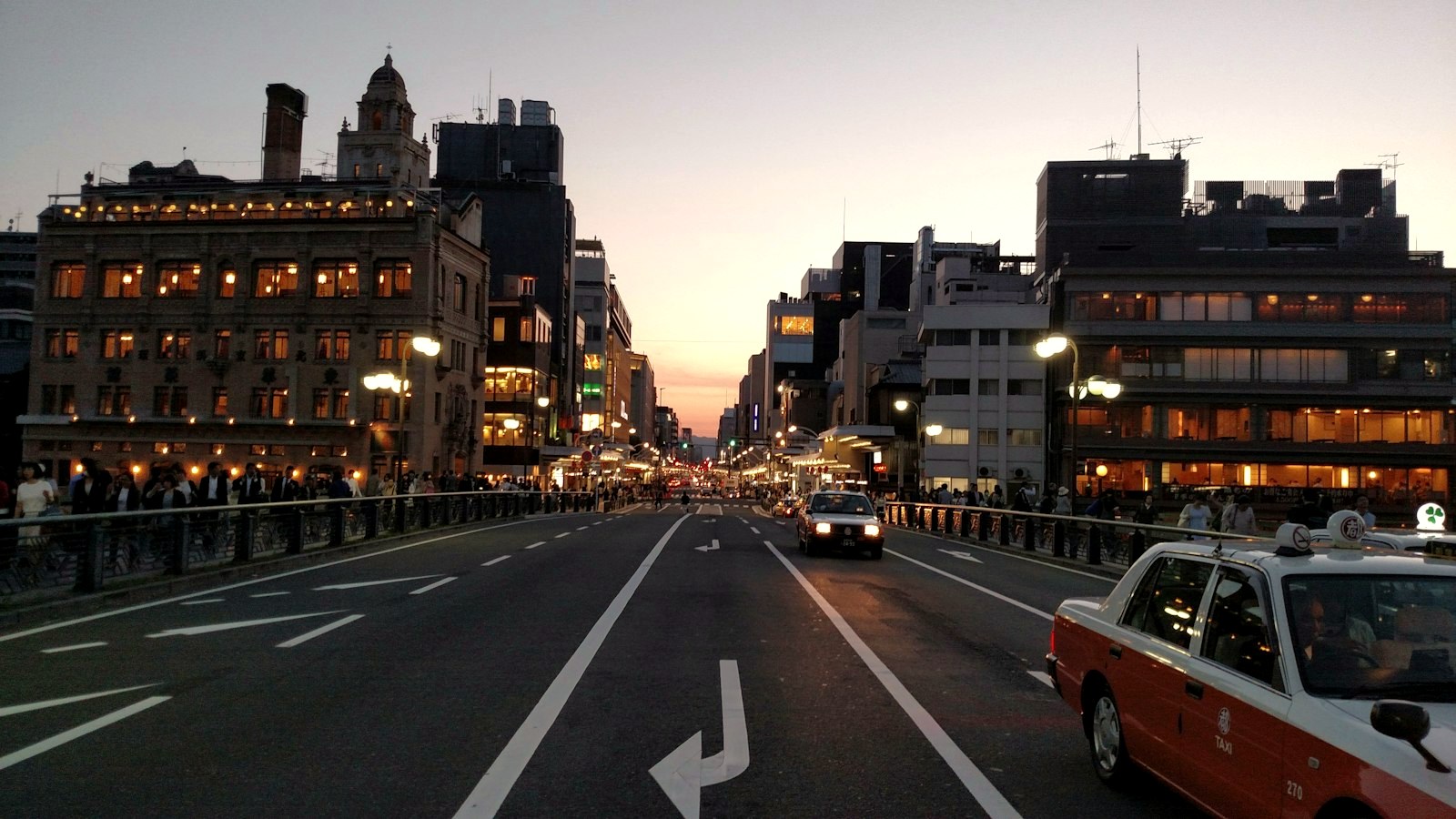 A bridge in Kyoto at dusk with a red taxi crossing, restaurant lights along the river