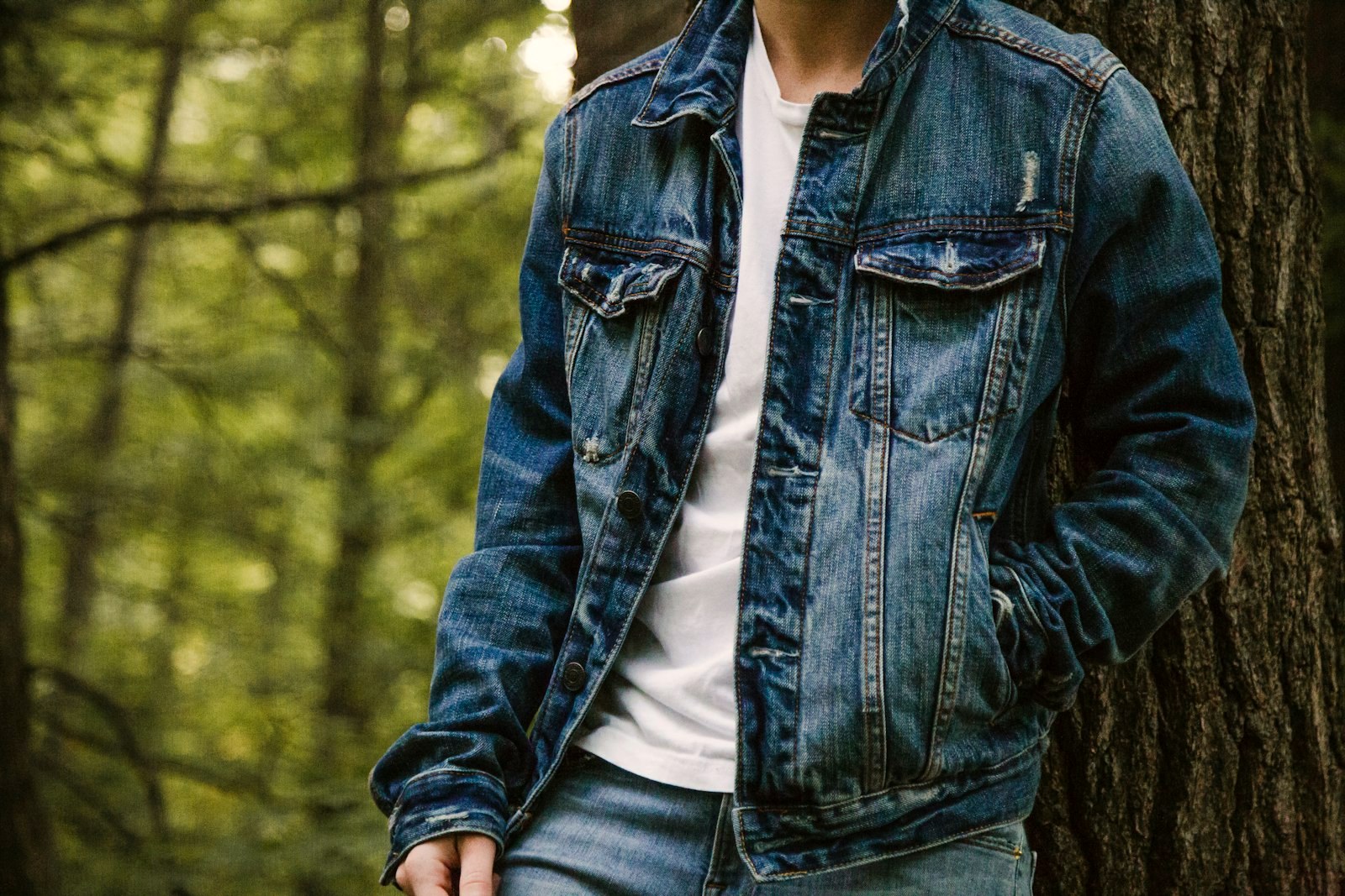 A man in a denim trucker jacket over a white t-shirt, standing in woodland light