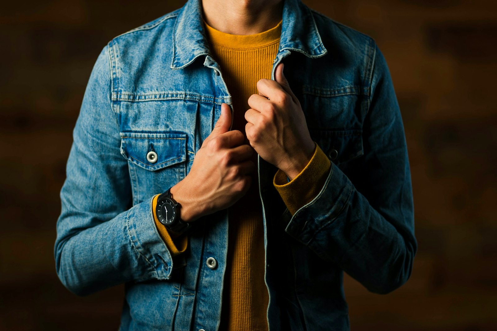 A man in a denim jacket over a mustard knit, hands at the placket