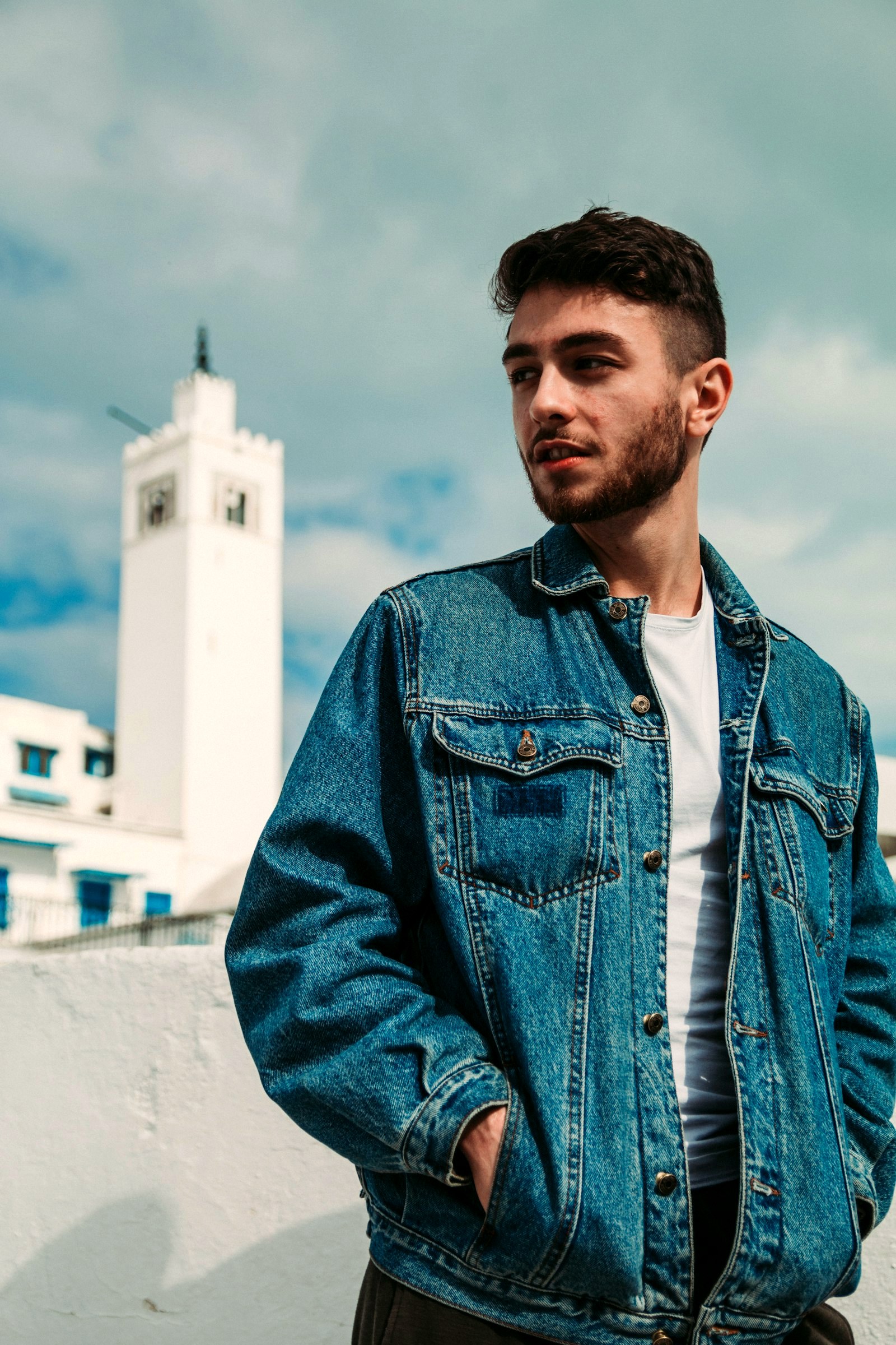 A man in an oversized denim jacket over a white t-shirt, a white tower behind him
