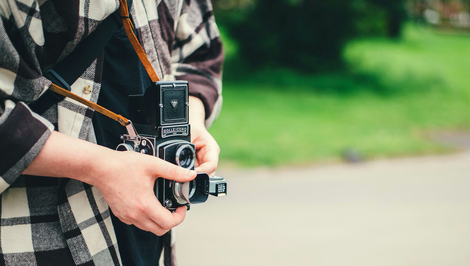 A man in a checked flannel shirt holding a vintage film camera, park light behind