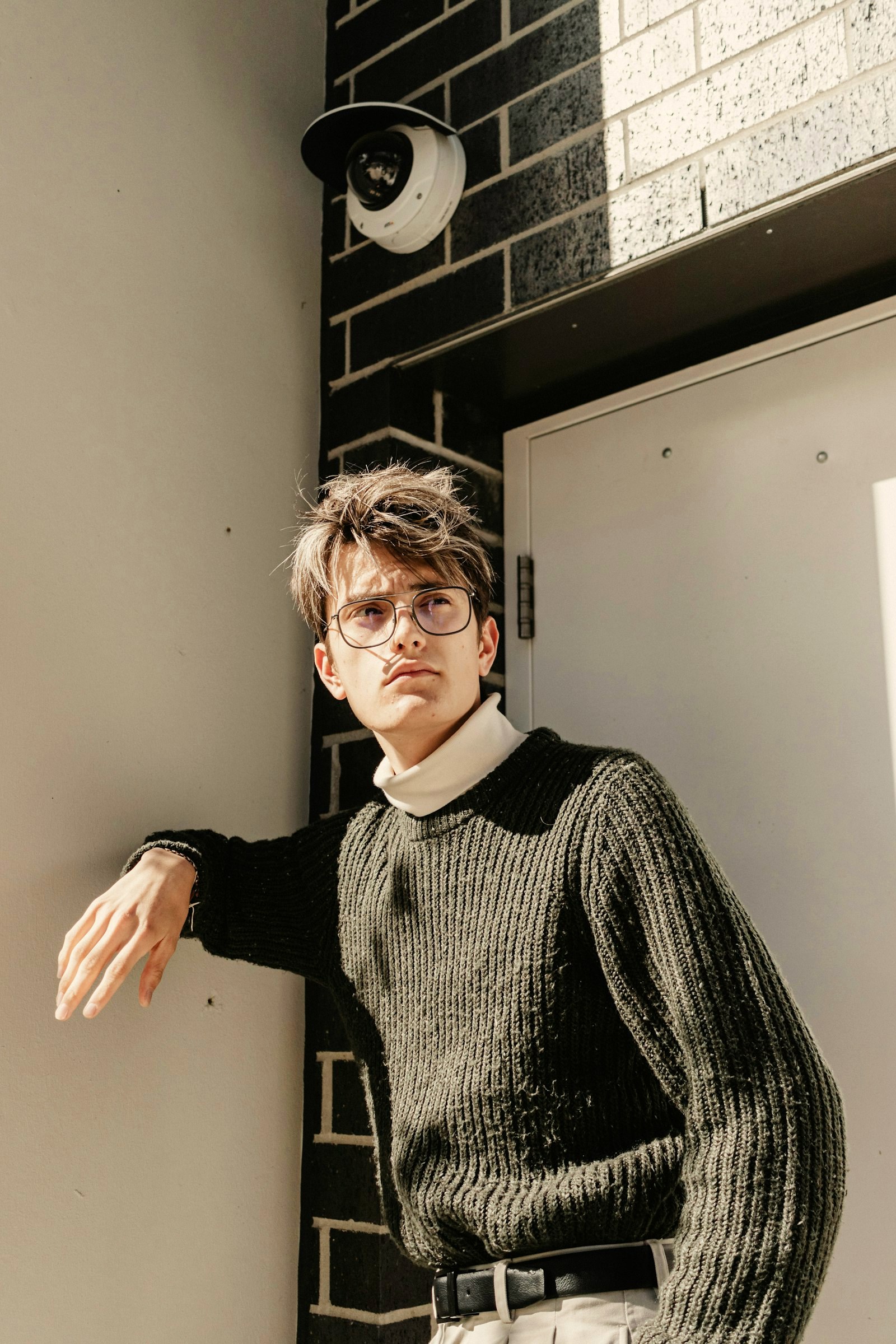 A man in a chunky ribbed knit turtleneck leaning against a tiled city wall