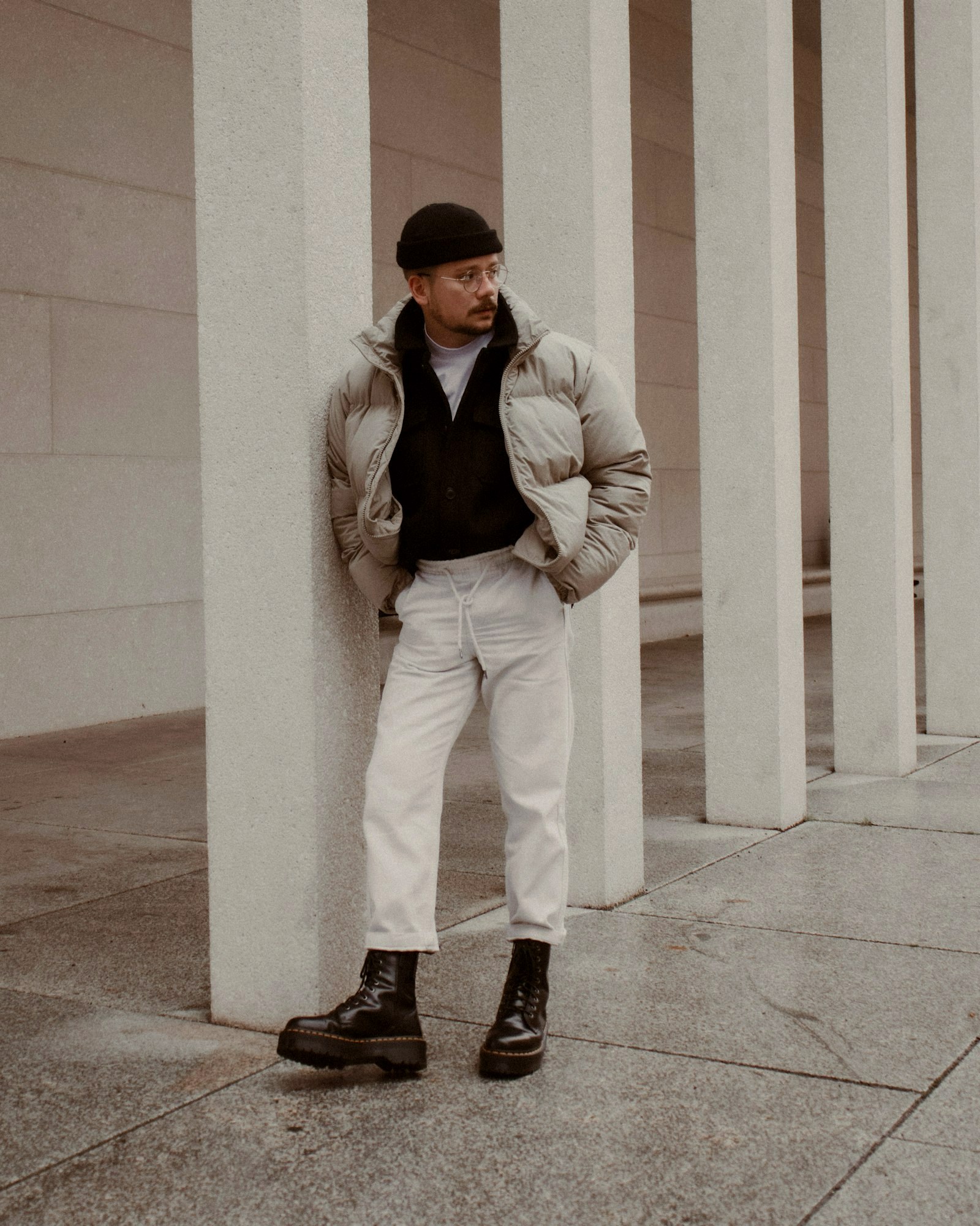 A man in a long pale puffer coat and white trousers leaning against white columns