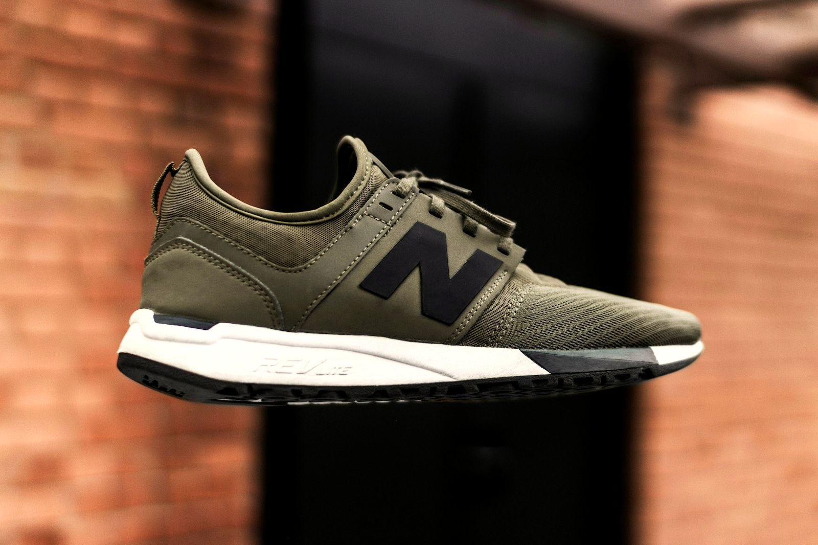 An olive-green New Balance sneaker floating against a brick wall
