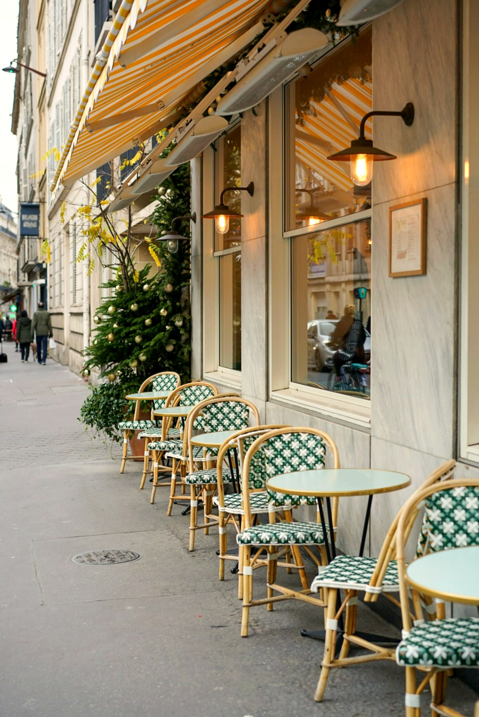 A row of rattan cafe chairs and small round tables under a striped awning, morning light