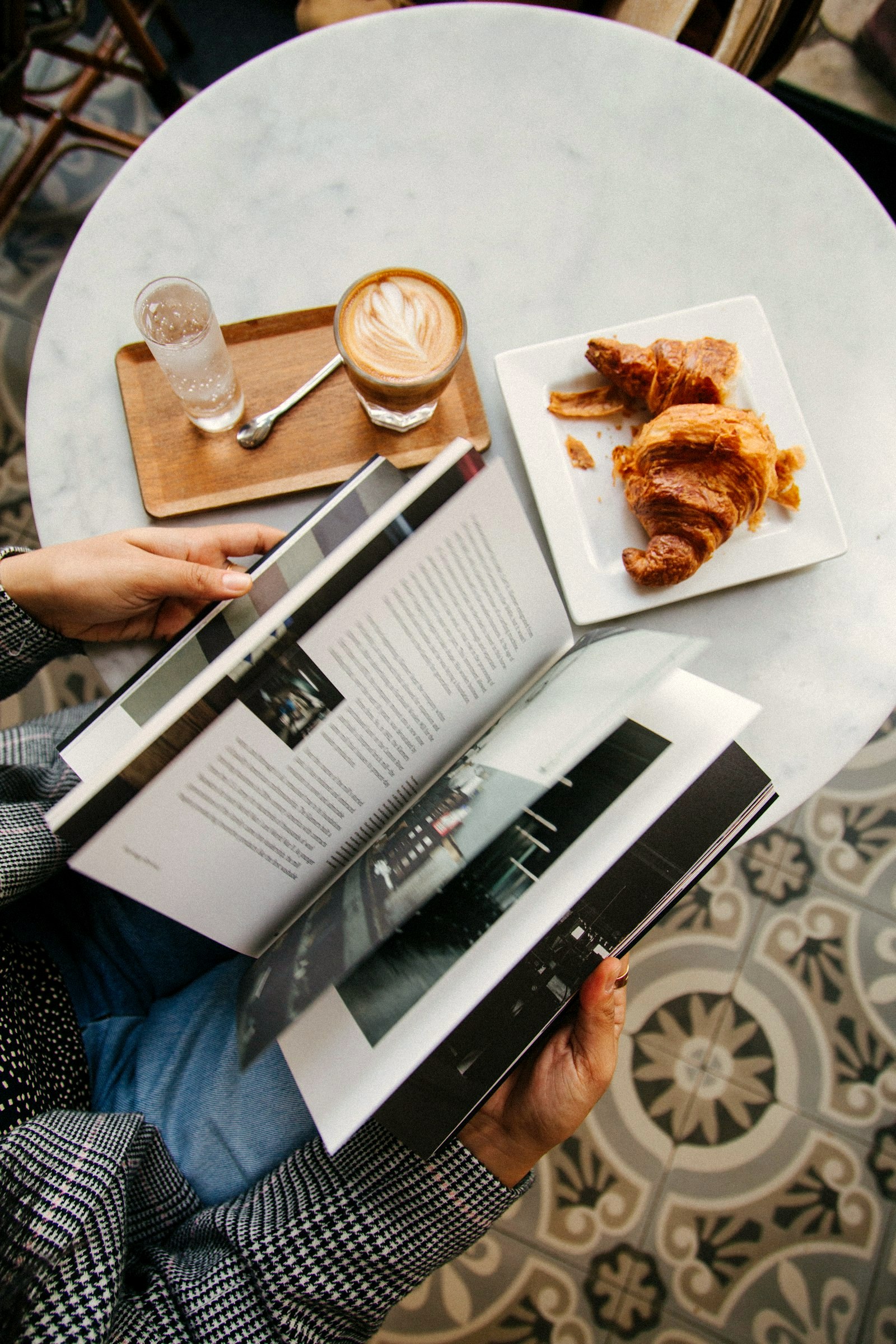 Overhead view of a cafe table: latte art, croissants and hands holding an open magazine