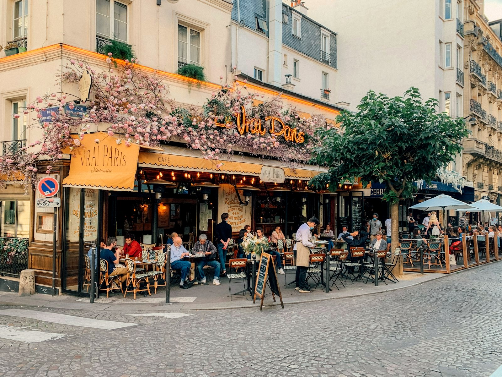 A Paris cafe corner with blossom over the terrace, diners at rattan tables, cobblestones