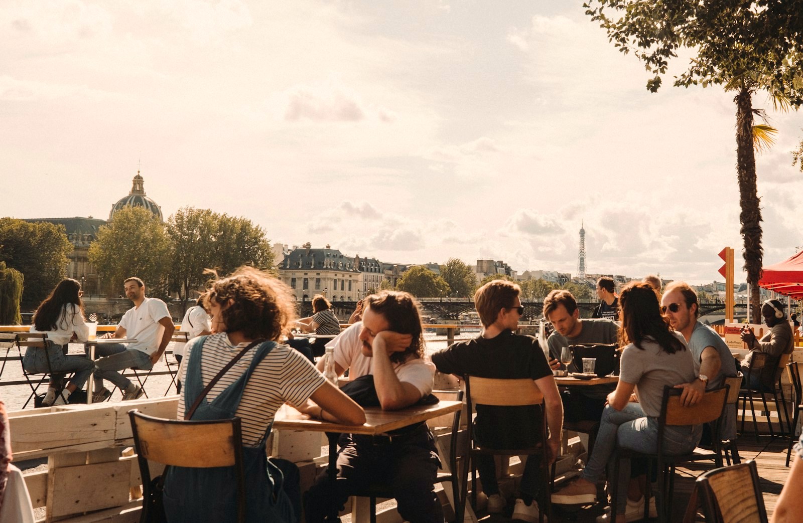 Diners at riverside cafe tables in golden hour, the Seine and a distant tower behind