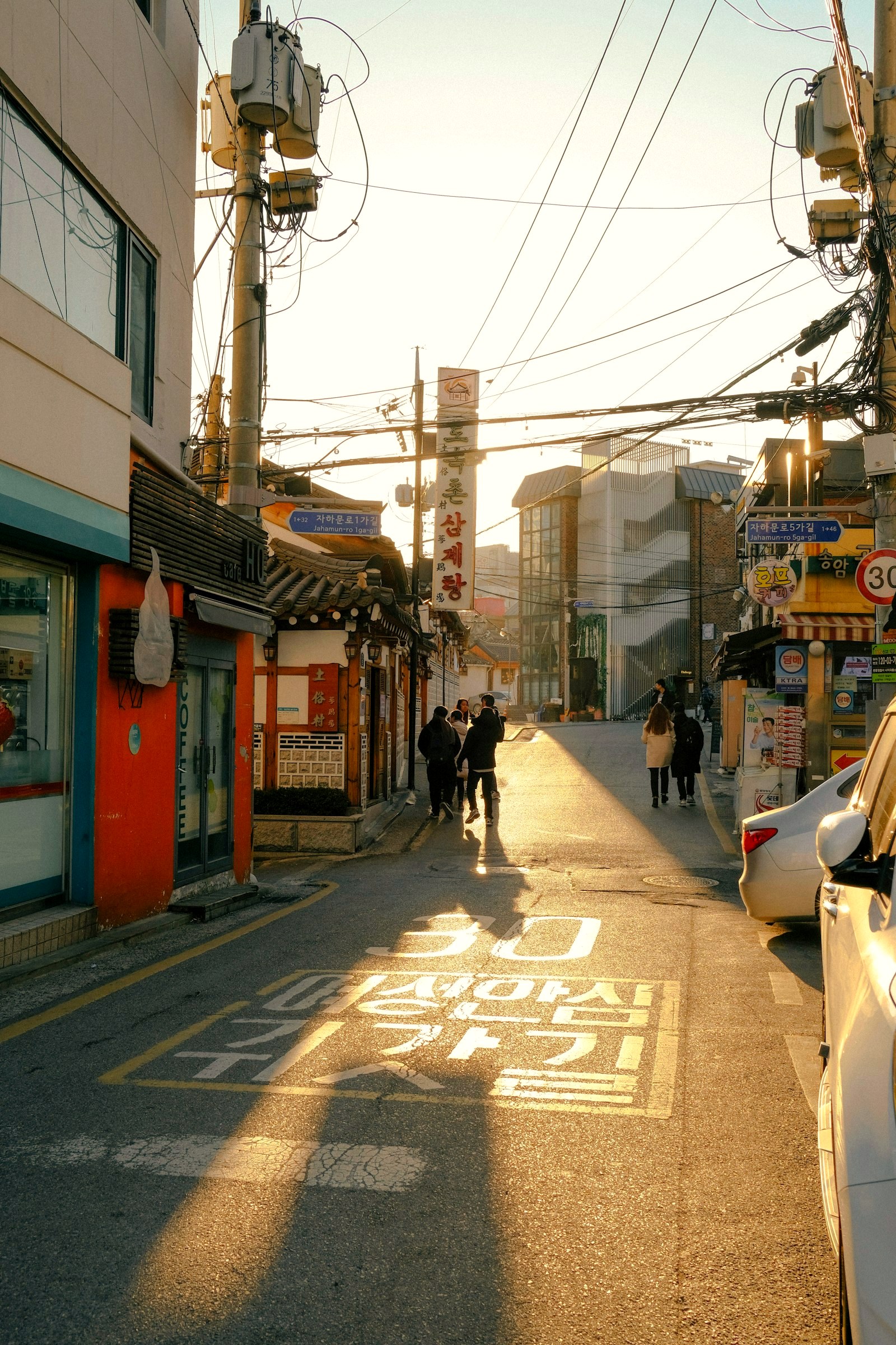 A quiet Seoul alley at golden hour, hanok rooflines beside modern storefronts, people in long coats walking away