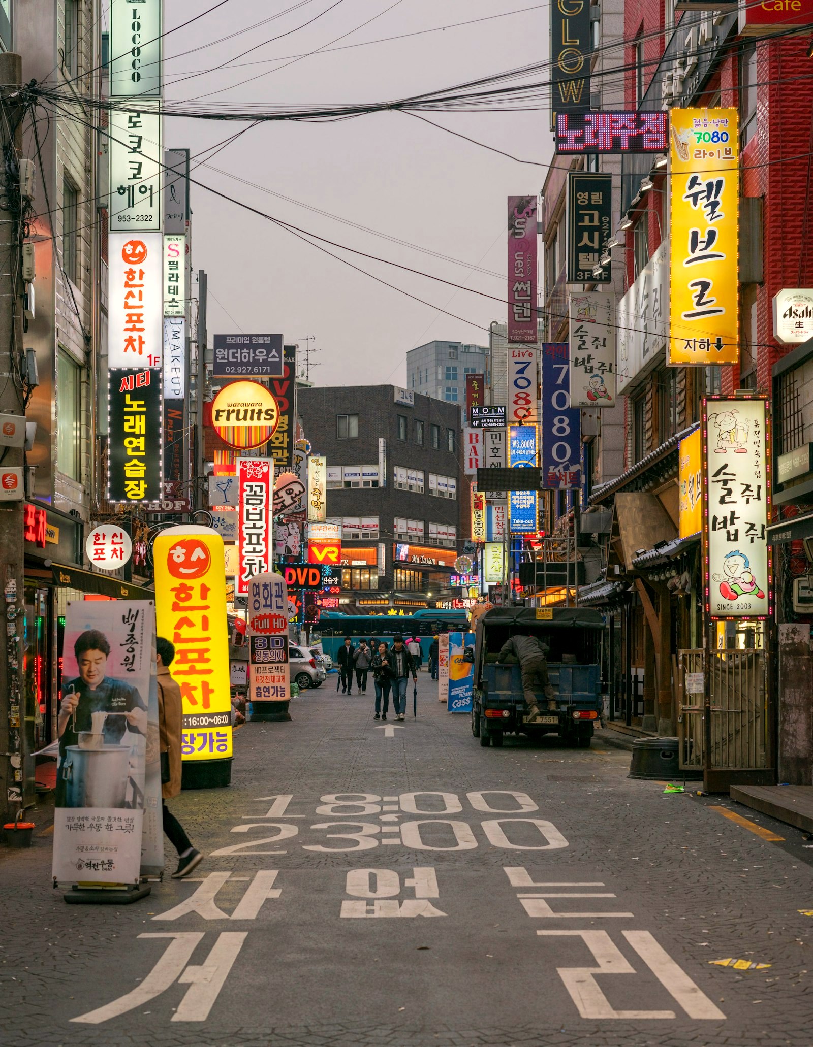 A shopping street at dusk under dense vertical signage, pedestrians crossing