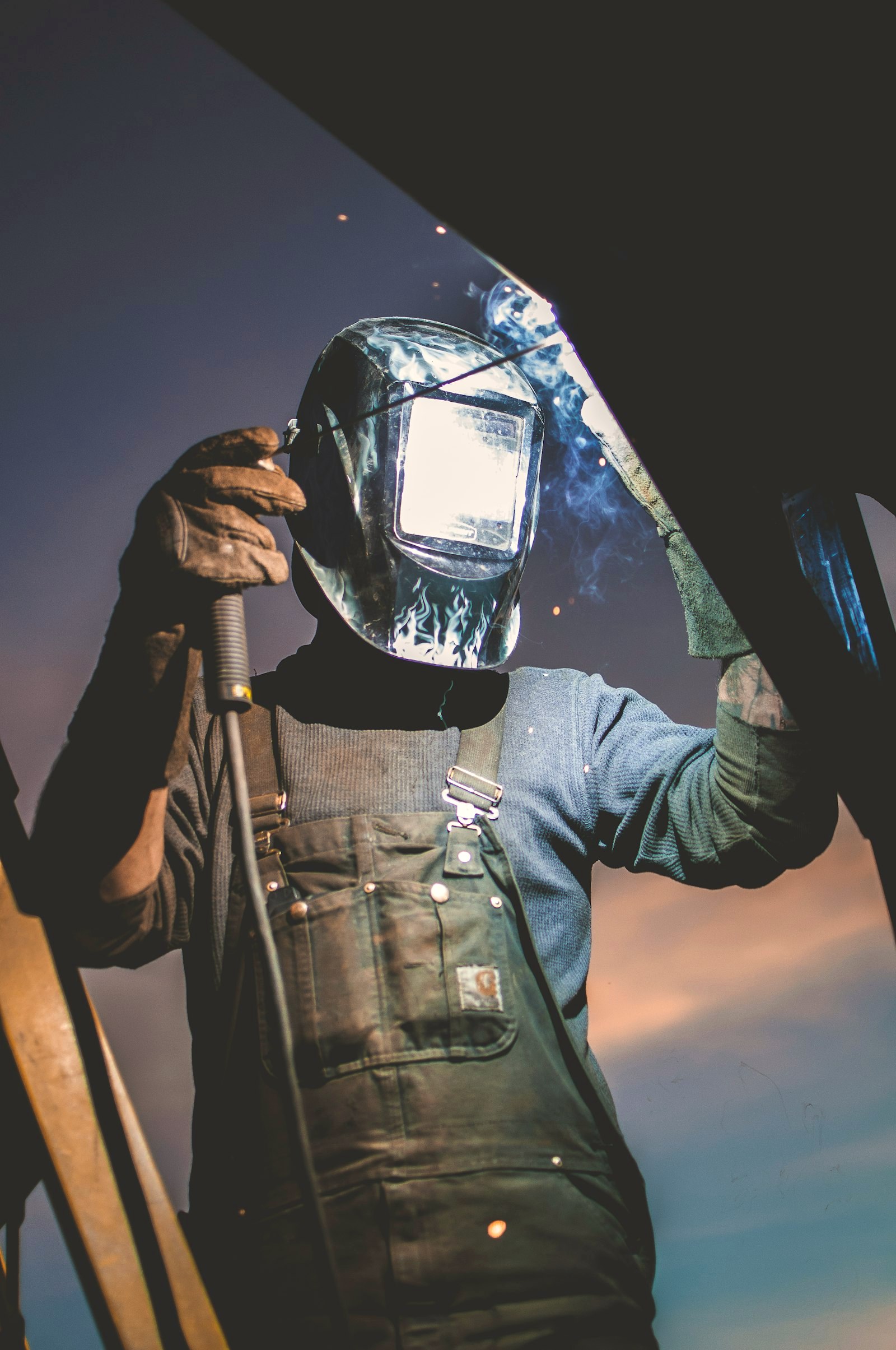 A welder in canvas overalls raising his mask mid-arc, sparks and smoke in dusk light