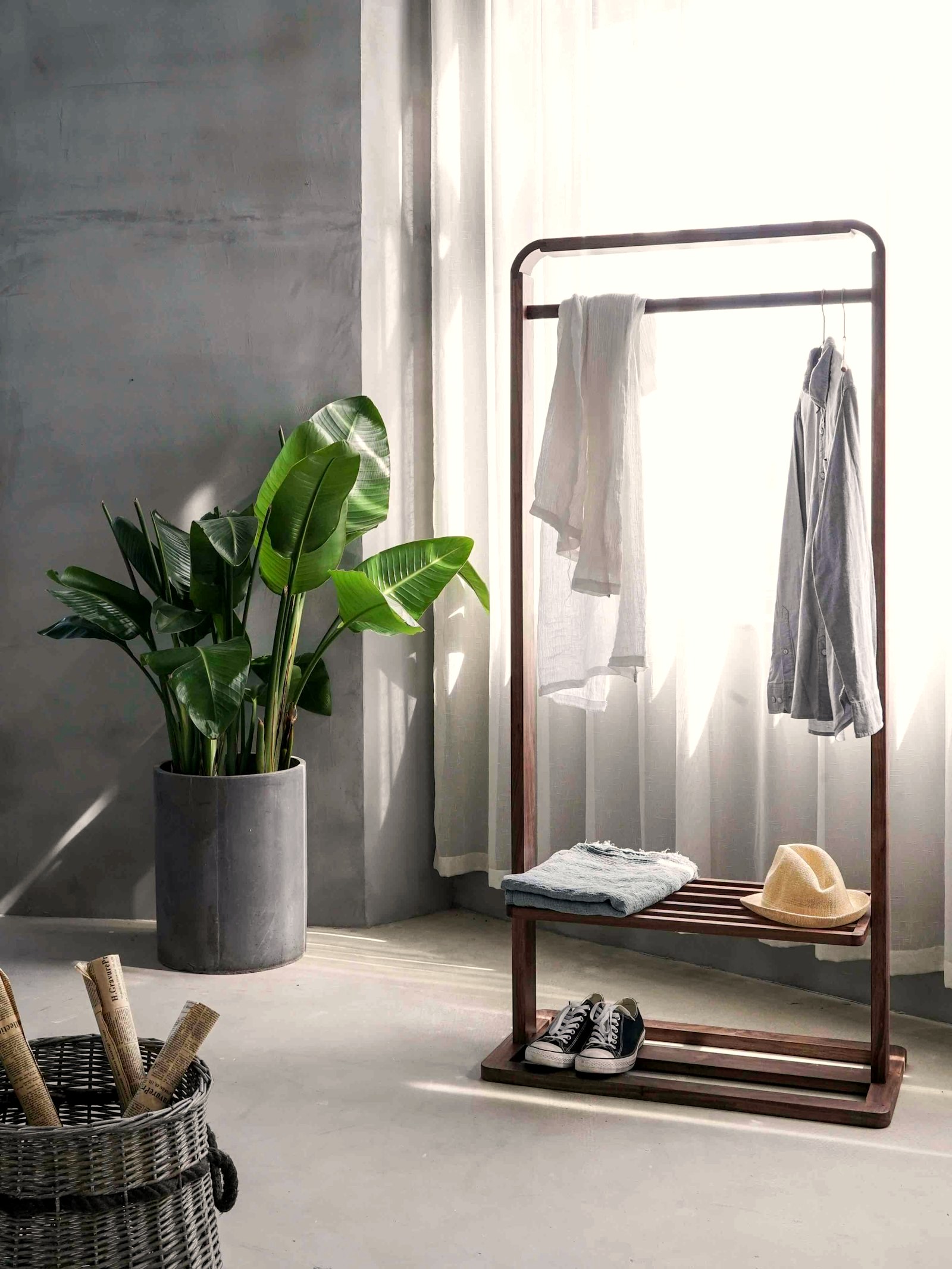 A wooden garment rail holding two shirts and a folded knit beside a bright window, sneakers on the shelf below
