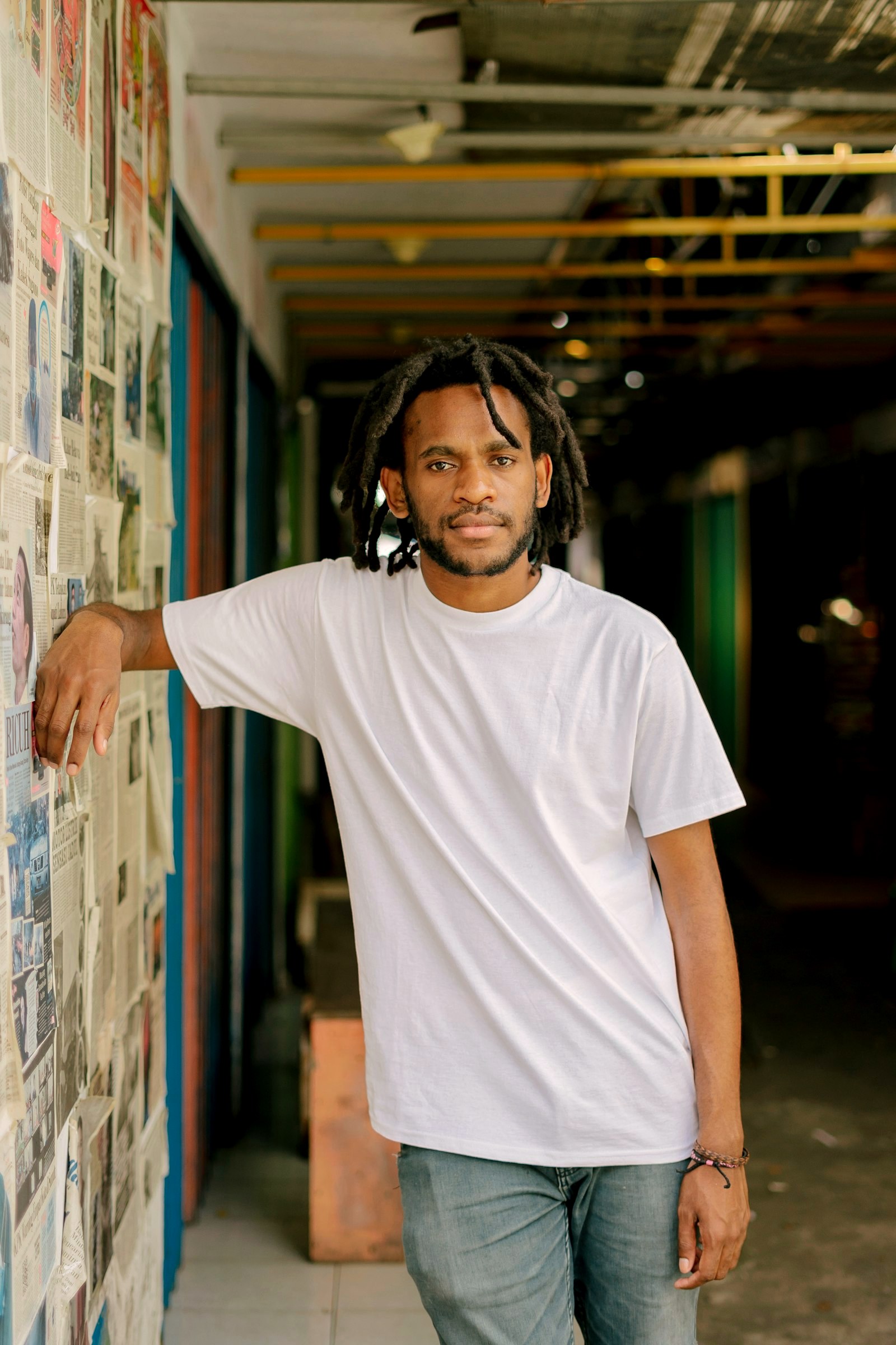 Young man with locs in a plain white t-shirt leaning in a market corridor